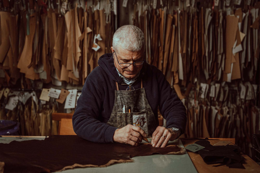 Experienced artisan working on leather at a workshop table, surrounded by hanging templates.