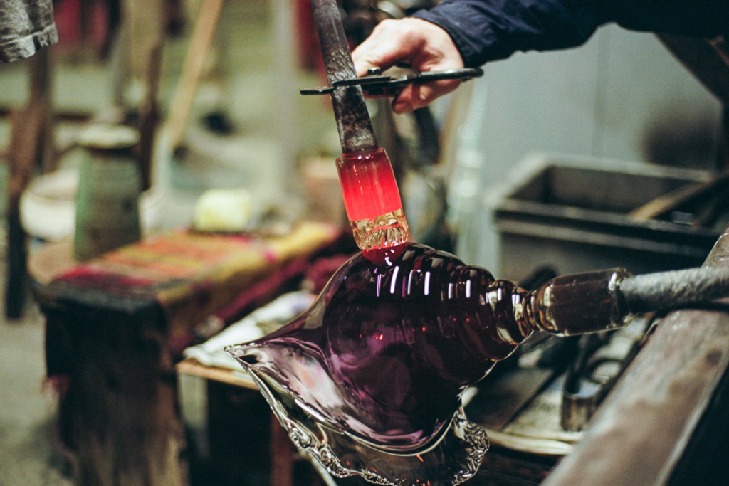 A glassblower shapes molten purple glass with tools, creating an intricate glass piece in a workshop setting.